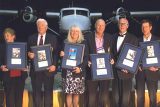 Six people standing in front of an airplane, each holding framed awards. They appear to be on a stage or platform. The airplane is large and prominently displayed in the background.