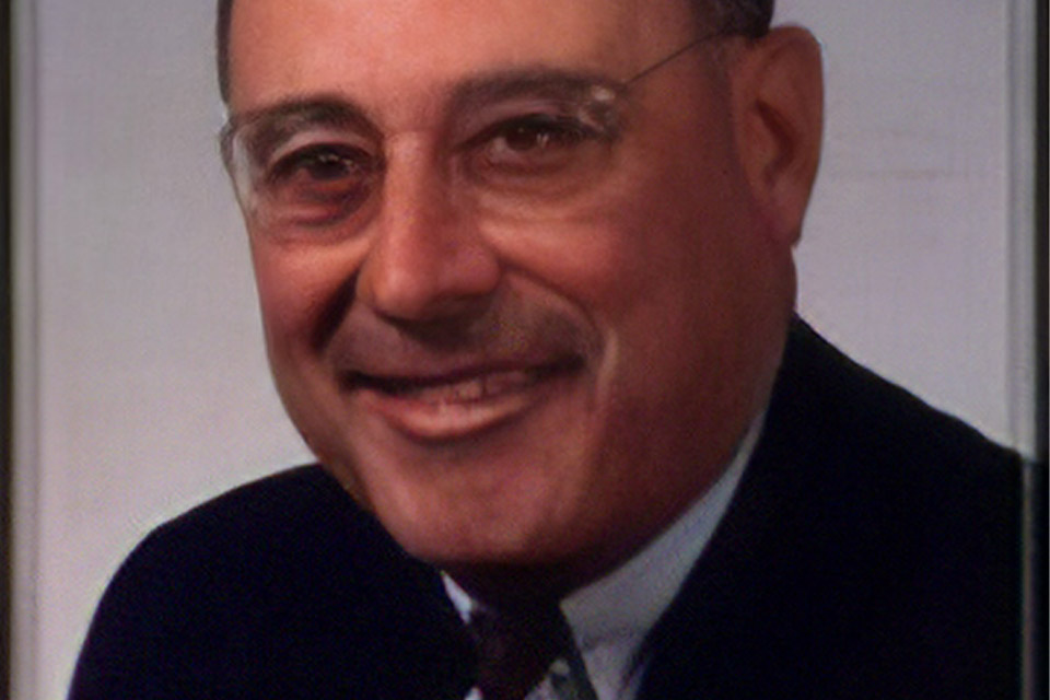 Smiling person with glasses wearing a suit and tie, against a plain background.