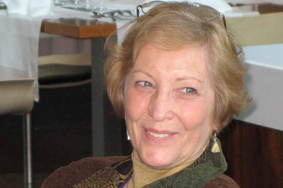 An older woman with short, light brown hair smiles warmly. She's wearing a brown sweater and a patterned scarf, with dangling earrings. The background shows a dining setting with tables and chairs.