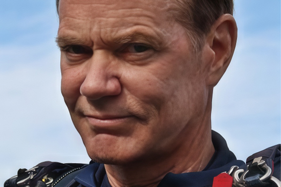 A close-up of a man with short hair, wearing a dark jacket, looking intently at the camera against a clear blue sky background.