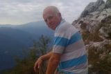 An older man with a shaved head, wearing a blue and white striped shirt, sits on a rocky cliffside. He looks toward the camera with mountains in the background under a cloudy sky.