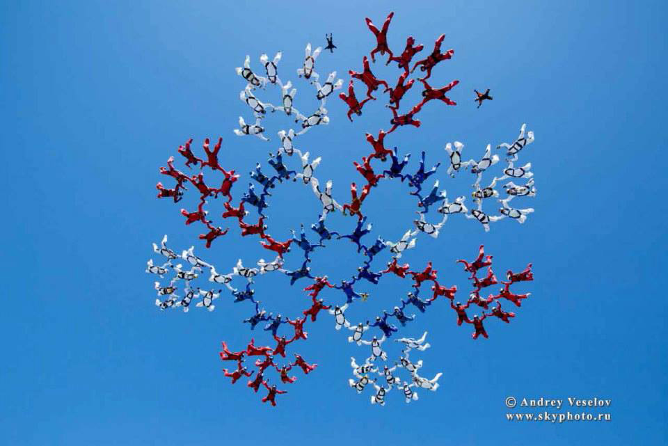 A large group of skydivers in red, white, and blue suits form a snowflake-like pattern against a clear blue sky. The formation is symmetrical and intricate, showcasing coordination and teamwork.