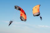 Two parachutists in mid-air with bright orange parachutes against a clear blue sky. They are approaching one another, with their parachutes partially open and lines visible. The scene conveys a sense of adventure and freedom.