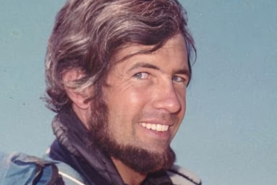 Smiling man with medium-length dark hair and a beard, wearing a blue jacket against a clear blue sky background.