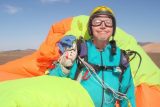 A person wearing a turquoise jumpsuit and helmet stands outdoors, smiling, holding bright orange and green parachute gear. The landscape is desert-like with mountains in the background under a clear blue sky.