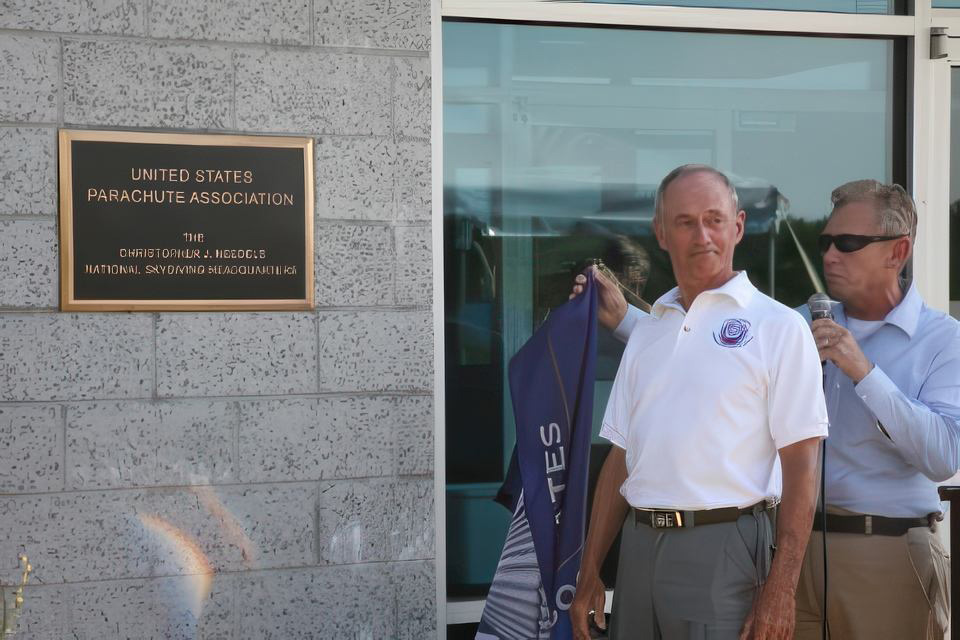 Two men standing in front of a building with a plaque reading "United States Parachute Association." One man holds a microphone while the other holds a folded flag. They appear to be attending a formal event.