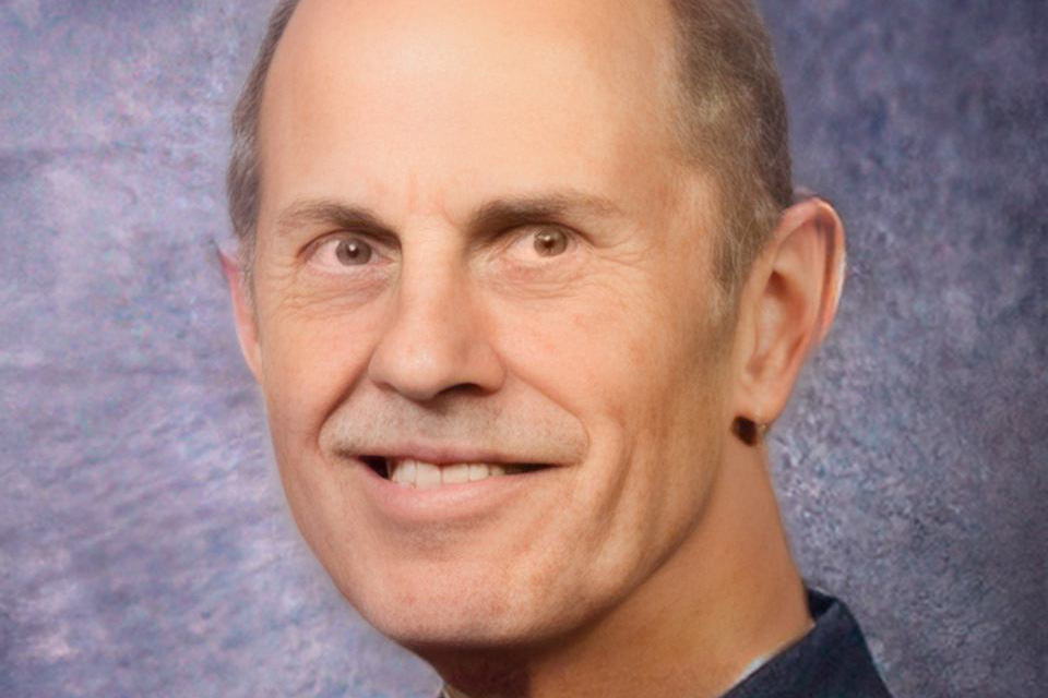 A mature man with short hair smiles at the camera against a textured blue-grey background. He is wearing a dark shirt.