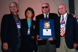 Four people stand together at an event. One of them holds a framed photograph with a portrait and accompanying text. They all wear event badges and formal attire. The background has a simple curtain.