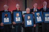 Five people holding framed awards with photos and text stand in a line, smiling. The background features a red curtain and a logo on a banner.