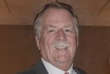 A man with gray hair and a mustache is smiling at the camera. He is wearing a suit and tie, and the background is softly blurred.