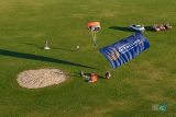 A skydiver is landing on a grassy field with a large blue banner reading "Skydiving Show and Challenge." Nearby, equipment and vehicles are visible, along with other people preparing for or observing the event.