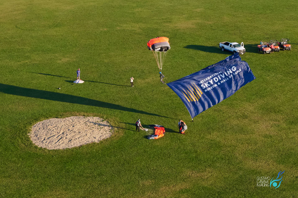 A skydiver is landing on a grassy field with a large blue banner reading "Skydiving Show and Challenge." Nearby, equipment and vehicles are visible, along with other people preparing for or observing the event.