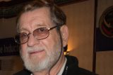 Elderly man with glasses and a gray beard, smiling slightly at the camera, wearing a black jacket. A softly lit indoor setting with a partial view of a decorative wall and a sign in the background.