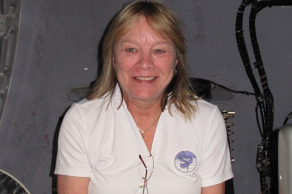 A woman with shoulder-length hair smiles while sitting indoors. She is wearing a white shirt with a logo and glasses hanging from her collar. The background shows some cables and equipment.