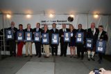 A group of people standing in a row at a Hall of Fame banquet, each holding a framed plaque. They are under a white tent with a sign that says "Hall of Fame Banquet and Induction Ceremony." The setting appears formal.