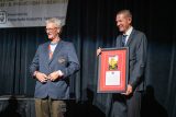 Two men stand on a stage. One, in a suit, holds a framed picture and plaque. The other, wearing a blazer with a patch, smiles. A banner in the background reads "Parachute Industry Association" at an event.