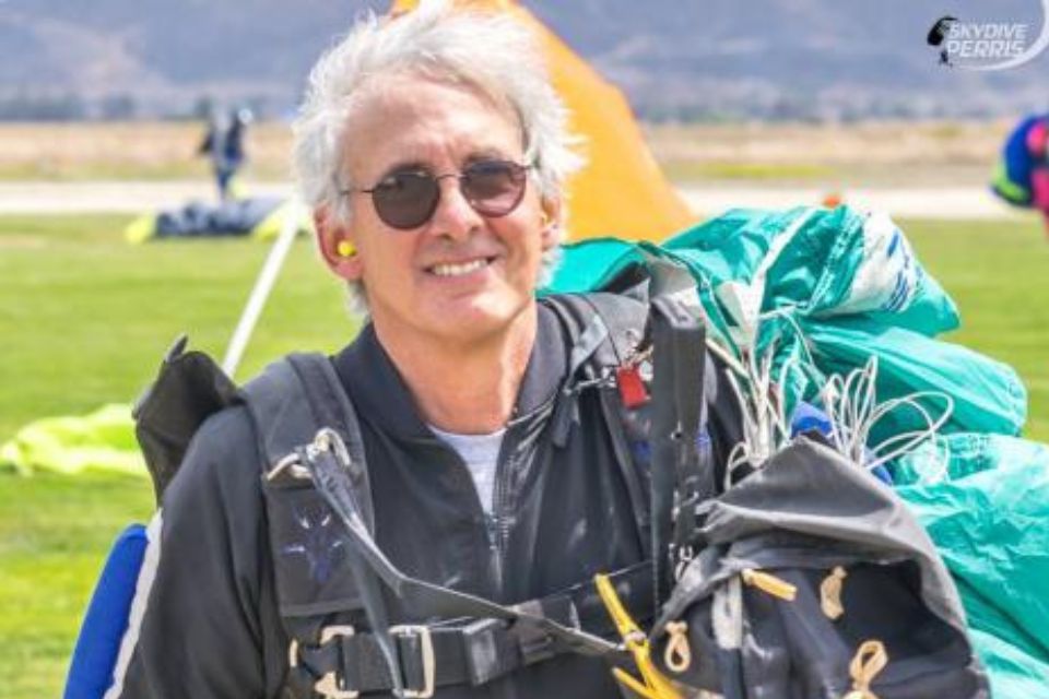 A person with white hair and sunglasses smiles while holding parachute equipment. They are wearing a dark jumpsuit and yellow ear protection, standing on a grassy field with a parachute canopy visible in the background.