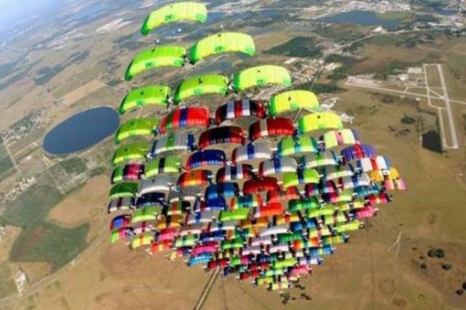 A large formation of skydivers with colorful parachutes is seen from above, creating a vibrant pattern against a landscape of fields and a nearby airport runway.