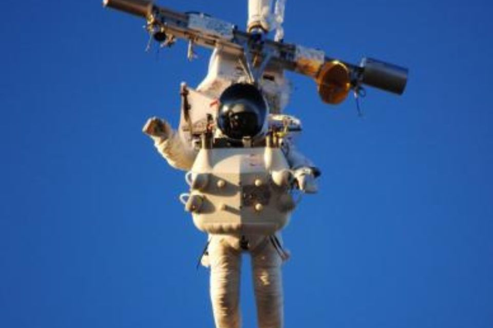 An astronaut in a white spacesuit performs a spacewalk against a clear blue sky, holding onto a large piece of equipment with various instruments attached.