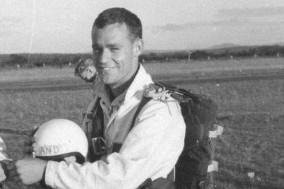 A skydiver in a jumpsuit smiles at the camera while holding a helmet. He is equipped with a parachute and standing in an open field with a distant view of trees and hills in the background. The image is in black and white.