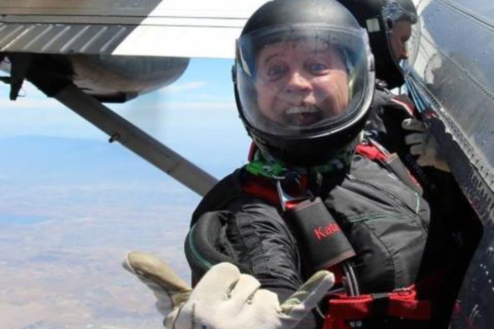 A person in a skydiving suit and helmet smiles excitedly while holding onto a plane door, preparing to jump. The background shows a clear sky and landscape below.