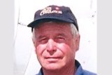 A middle-aged man wearing a dark baseball cap and a navy blue shirt is looking at the camera. The background is a plain, light-colored surface.