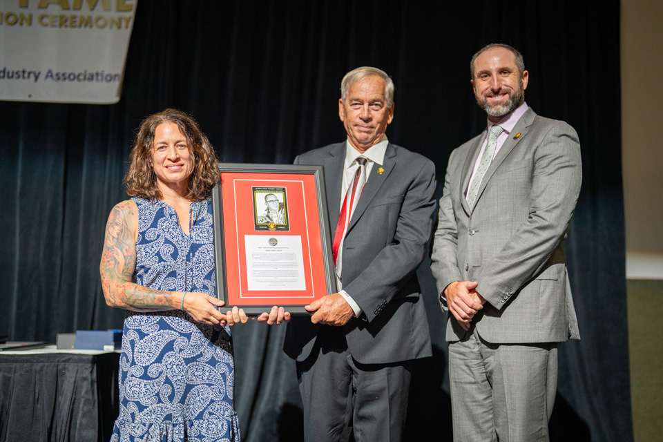 Three people pose on stage during an award ceremony. The person in the middle holds a framed award. A banner in the background reads "Induction Ceremony." They are dressed in formal attire, and the event appears celebratory.