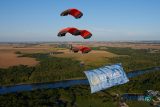 Four skydivers in formation with red parachutes descend over a scenic landscape of fields and a river. They are displaying a large banner that reads "International Skydiving Museum & Hall of Fame.