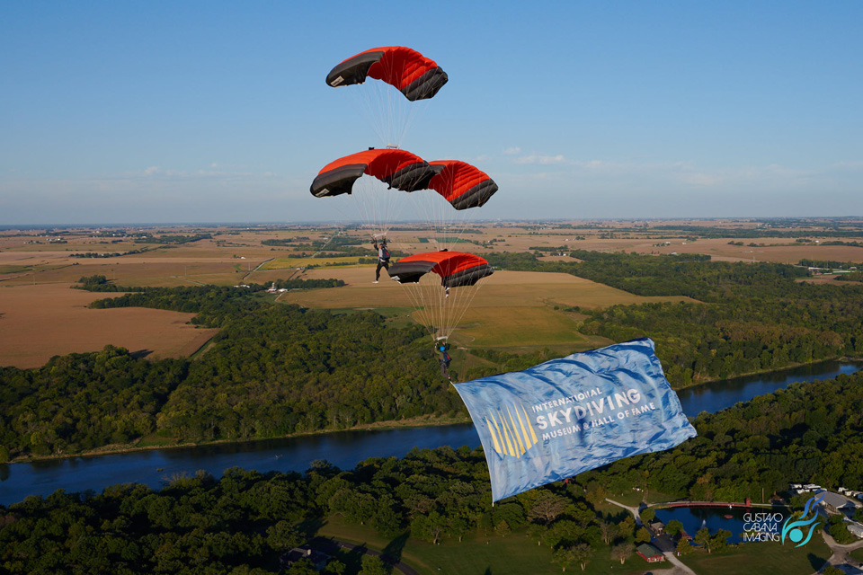 Four skydivers in formation with red parachutes descend over a scenic landscape of fields and a river. They are displaying a large banner that reads "International Skydiving Museum & Hall of Fame.