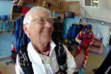 An older woman with white hair smiles while wearing a white skydiving jumpsuit and harness indoors. Behind her, another person in a brightly colored suit adjusts equipment. Shelves with helmets and gear are visible in the background.