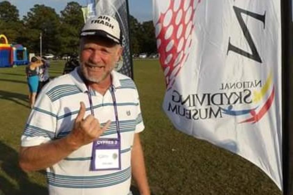 A man wearing a striped polo and cap stands on a grassy field, making a "hang loose" hand gesture. He's near a flag with the text "National Skydiving Museum." People and trees are visible in the background under clear skies.