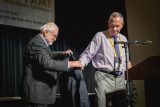 Two men on a stage during an induction ceremony. The man on the left is helping the man on the right put on a blazer. There's a microphone stand nearby and a banner partially visible in the background.