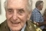 An elderly man wearing a military uniform smiles at the camera. Another person stands in the blurred background.