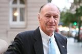 An older man in a suit and tie stands outdoors near a building, looking at the camera. The background features blurred city lights and a window.