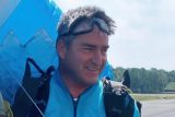 A man in blue skydiving gear stands outdoors, smiling. He wears goggles on his head and carries a parachute pack. Trees and a clear sky are in the background.