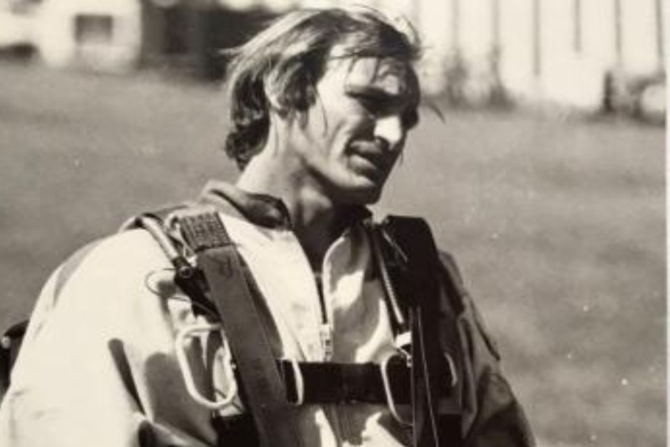 A black-and-white photo of a person in a parachute harness. They have short hair and are looking to the side. The background is a grassy field with a building out of focus.