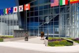 A modern building with a glass facade reflecting an airplane. Flags from various countries, including the US, South Korea, and Canada, are displayed above. Two people are reading a memorial with engraved names, surrounded by greenery and purple flowers.