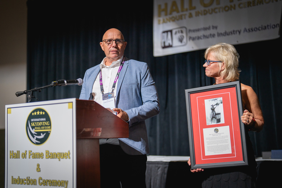 A man speaks at a podium during a Hall of Fame banquet, while a woman beside him holds a framed photograph and certificate. A banner in the background reads, "Hall of Fame Banquet & Induction Ceremony.