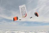 Two skydivers with orange and black parachutes fly through the sky, holding a large banner that reads "International Skydiving Museum & Hall of Fame." They are above fluffy white clouds.