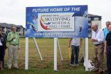 A group of people stand around a sign that reads "Future Home of the International Skydiving Museum & Hall of Fame" with colorful skydiving images. The website www.SkydivingMuseum.org is displayed. The setting is a grassy field near buildings.