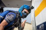 A person wearing a blue helmet with a camera, sunglasses, and skydiving gear gives a thumbs up while standing by an aircraft door. The person appears ready for a skydive and is smiling.