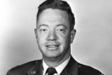 A black and white portrait of a man in a military uniform. He has short hair and is facing the camera with a slight smile. The uniform displays insignia and a tie. The background is plain.