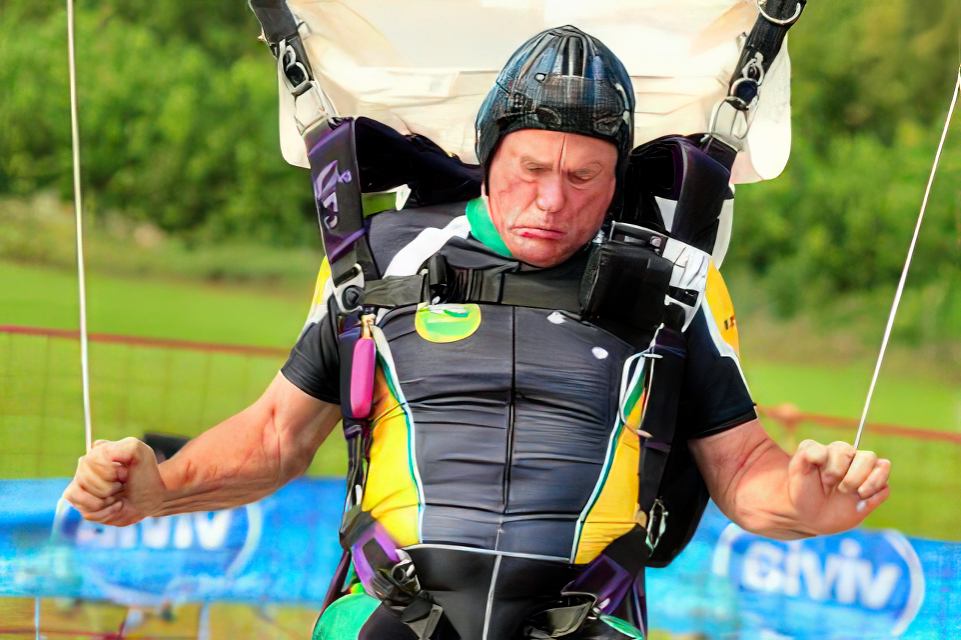 A person with an intense expression lands after skydiving, wearing a brightly colored jumpsuit and a helmet. Lush greenery is visible in the background.