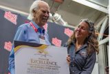 An older man in a blue shirt holds a large certificate titled "Path of Excellence." A woman in a dark shirt stands beside him, smiling. They are indoors, with a "Skydive Arizona" logo visible in the background.