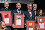 A group of six smiling people stand together, each holding a framed certificate. They appear to be at an award ceremony or event, with a banner visible in the background.