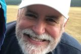 A smiling man with a white beard and a white cap, wearing a dark shirt, stands outdoor in a field. The background is blurred, showing a mix of grass and trees.