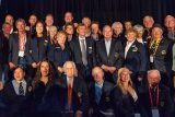 A group of people, mostly older adults, pose together wearing dark blazers with emblem patches. They are smiling in front of a dark curtain backdrop. Some are seated while others stand behind them.