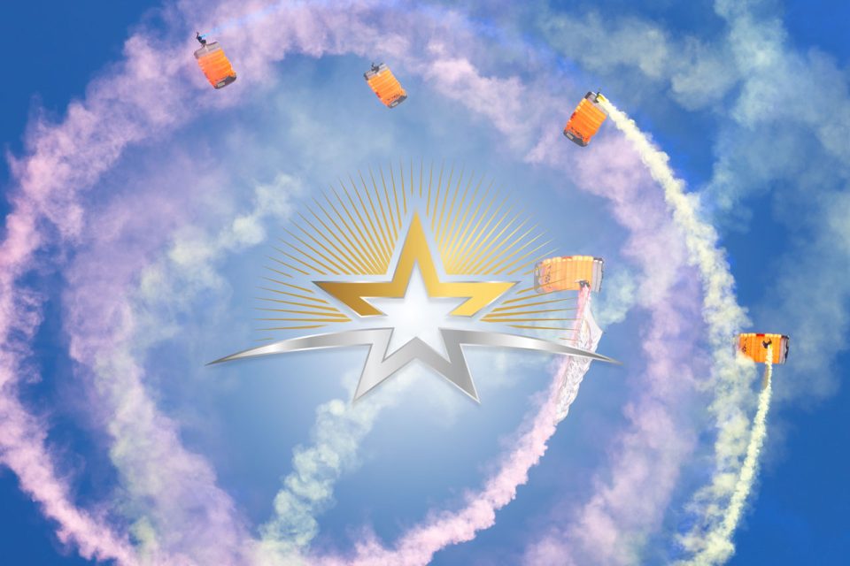 Skydivers with colorful smoke trails are descending in formation against a blue sky. A large, stylized silver and gold star with an aura is superimposed in the center of the image, radiating streaks outward.