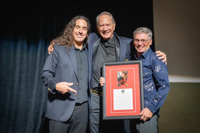 Three men are standing together, smiling. The man in the center is holding a framed certificate or award. They are dressed in suits, and a dark curtain is in the background.