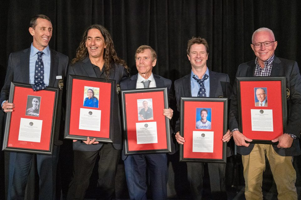 Five men stand side by side, each holding a framed certificate with a photo, against a dark background. They are dressed in formal attire and smiling at the camera.